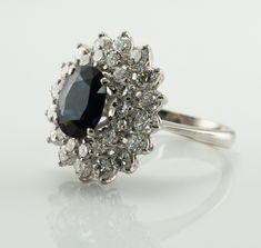 This estate ring is crafted in solid 18K White Gold (carefully tested and guaranteed). The center natural Earth mined oval cut Sapphire measures 9x7mm (2.20 carats). This gem has a deep blue color. It also has a tiny chip that cannot be seen without magnification. The center stone is surrounded with 32 round brilliant cut diamonds. 16 diamonds total 1.28 carats and smaller 16 gems total .64 carats. The grand total diamond weight is 1.92 carats of VS1 clarity and G color. Size 7 (sizable). The ri Formal Diamond Flower Ring With Gemstone, Elegant Flower Ring With Polished Finish For Anniversary, Elegant Formal Flower Ring With Diamond Cut, Luxury Oval Cluster Ring With Diamond Cut, Heirloom Cluster Ring With Polished Finish, Oval Shape, Heirloom Cluster Ring With Polished Finish, Timeless Oval Cluster Ring Hallmarked, Elegant Hallmarked Cluster Ring Marquise Cut, Elegant Oval Cabochon Sapphire Wedding Ring