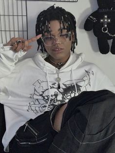 a young man with dreadlocks wearing a white hoodie and black pants sitting in front of a teddy bear