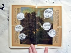 a hand holding an open book with trees and speech bubbles above it on a white surface