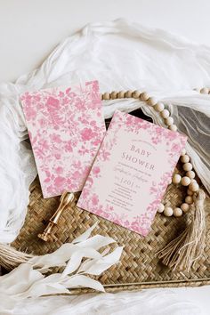 two pink and white baby shower cards sitting on top of a wicker basket next to beads