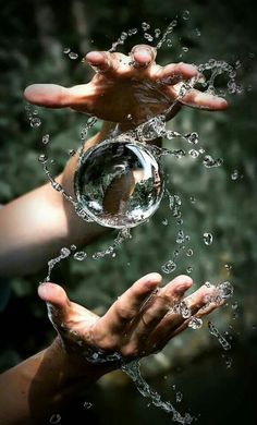 two hands are holding a glass ball with water splashing out of it to the camera