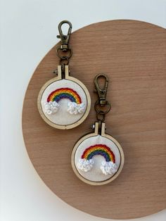 two key chains with embroidered rainbows and clouds on them sitting on a wooden surface