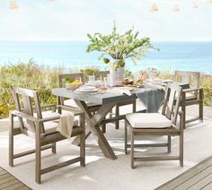 an outdoor dining table set with chairs and plates on it, overlooking the beach in the background