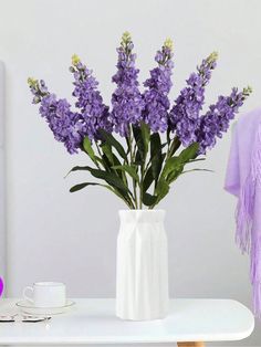 a white vase filled with purple flowers on top of a table next to a cup