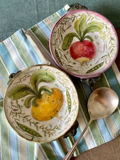 two bowls with fruit painted on them are next to spoons and a striped napkin