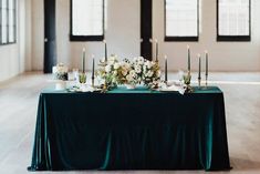 a long table with candles and flowers on it in front of two windows, all set up for a formal dinner