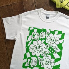 "Grow With The Flow Graphic T-Shirt - Oversized Unisex White Tee - 100% Cotton, Eco-Friendly - Get Gardening In Style White cotton tee illustrated with a green graphic print of smiley flowers, wavey checkerboard pattern and a cheery quote of \"Grow With The Flow\".  🌻 unisex t-shirt 🧵 100% cotton 📏 relaxed fit ⚖ midweight fabric 203 gsm 🌍 planet friendly 💛 sustainably sourced materials  💧 printed using vegan friendly, water-based inks  🌟 made to order, printed just for you! (a more sustainable method that avoids unnecessary textile waste) 🐳 posted in a plastic free biodegradable mailing bag ♻ packaging is 100% recyclable after use ©2024 Kio Cards Ltd. All images, artwork and content. All rights reserved. say hi/tag us on Instagram @sixsunflowersdesign" Relaxed Fit Green T-shirt With Plant Print, Oversized Green Graphic T-shirt, Green Cotton T-shirt With Plant Print, Spring Cotton Shirt With Graphic Design, Green T-shirt With Plants Print, Relaxed Fit, Green Shirt With Text Print For Spring, Green Cotton Tops With Graphic Design, Green Cotton Shirt With Plant Print, Oversized Green Graphic Tee Shirt