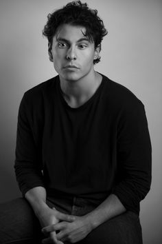 a man sitting down with his hands on his knees and looking at the camera, in black and white