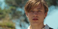 a close up of a person wearing a white shirt and looking at the camera with trees in the background