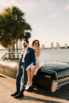 a man and woman sitting on the hood of a black car in front of a body of water