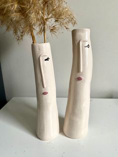 two white vases with faces on them sitting next to a plant in front of a wall