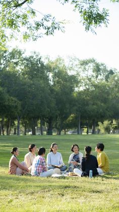 a group of people sitting on top of a lush green field under a tree filled sky