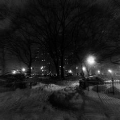 a black and white photo of snow on the ground at night with street lights in the distance