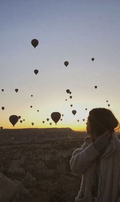 a woman standing on top of a field with lots of hot air balloons in the sky