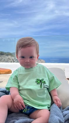 a little boy sitting on top of a bed next to the ocean and wearing a green shirt