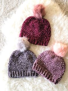 three knitted hats laying on top of a fluffy white rug with pom - poms