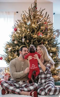 a man and woman sitting in front of a christmas tree holding a baby under it