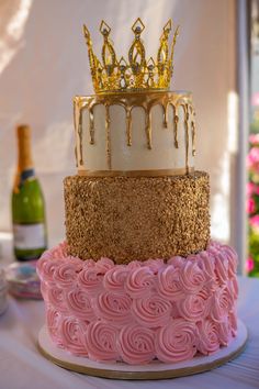 a three tiered cake with pink icing and gold crown on top, sitting on a table