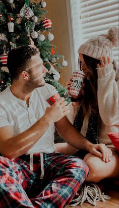 a man and woman sitting on the floor next to a christmas tree drinking coffee together