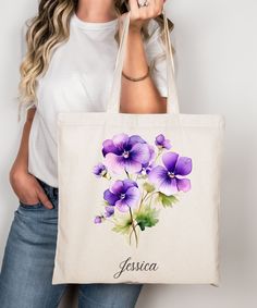 a woman holding a tote bag with purple pansies on it and the word fossia written in cursive writing