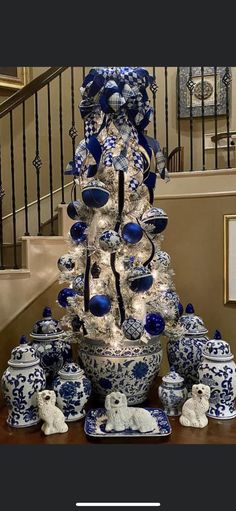 a christmas tree with blue and white ornaments on it in front of stairs, next to a staircase case