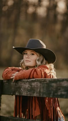 a woman wearing a cowboy hat leaning on a wooden fence with her arms crossed and looking off to the side