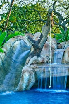 an artificial waterfall in the middle of a pool surrounded by trees and rocks with a rope hanging from it