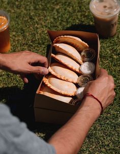 a man is opening a box of food on the grass next to two cups of coffee