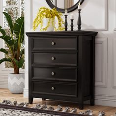 a black dresser with three drawers and a round mirror on the wall above it, next to a potted plant