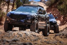 two trucks driving down a dirt road in the desert with rocks and trees behind them