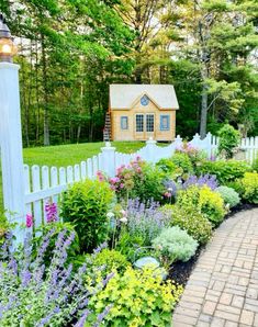 a white picket fence surrounded by colorful flowers and greenery in front of a small house