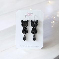 Black cat earrings with glittery details, perfect for Halloween or spooky-themed outfits. These cute and whimsical earrings are crafted from high-quality acrylic with a fun, dangling tail design that adds movement and sparkle. Lightweight and comfortable, they make a great accessory for Halloween parties or everyday spooky style. Ideal for cat lovers or as a festive gift, these black glitter cat earrings bring a touch of charm to any outfit.  Made with stainless steel findings for sensitive ears Halloween Cat Earrings, Cute Cat Design Earrings For Party, Party Jewelry With Cat Design And Cat Ears, Party Jewelry With Cat Design, Black Cat Ears Earrings For Party, Cute Black Party Earrings, Whimsical Earrings, Clay Halloween, Polymer Clay Halloween