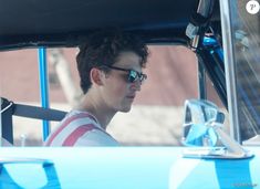 a young man driving a blue car with his sunglasses on and looking at something in the distance