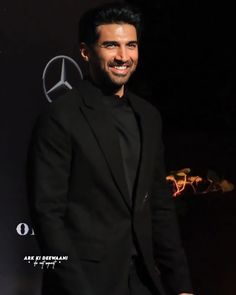 a man in a black suit smiling at the camera with his hand on his hip