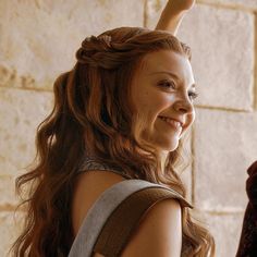 a woman with long hair is smiling and holding her hand up in the air while standing next to a brick wall