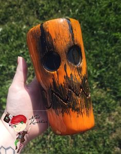 a hand holding up a wooden mask in front of some green grass with the face of a creature painted on it