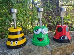 three different colored lamps with faces on them sitting on top of a rock near some bushes