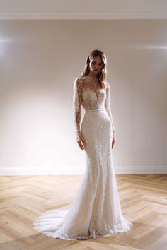 a woman in a long sleeved wedding dress standing on a wooden floor with her arms behind her back