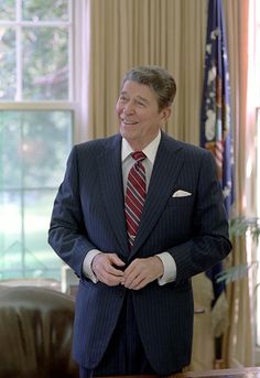 a man in a suit and tie standing next to a desk with an american flag behind him