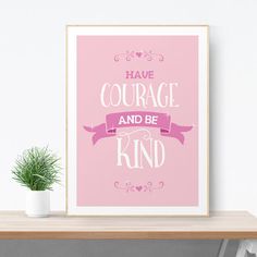 a pink poster with the words have courage and be kind on it next to a potted plant