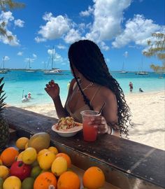 a woman sitting at a table with fruit and drinks in front of her on the beach