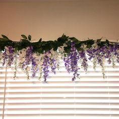 purple and white flowers hanging from the side of a window sill in front of blinds