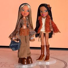 two dolls standing next to each other in front of a pink wall, one holding a purse and the other wearing brown boots