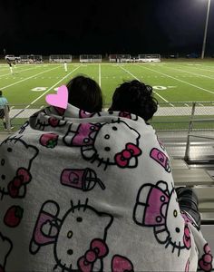 two people wrapped in hello kitty blankets on a football field at night with the lights on