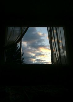 an open window with curtains and the sun setting in the sky behind it on a cloudy day