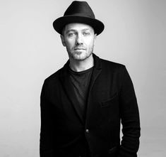 a man wearing a black hat and jacket standing in front of a white wall with his hands in his pockets