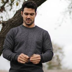 a man standing in front of a tree looking at his cell phone while wearing a gray sweater