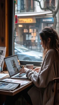 a woman sitting at a table with a laptop computer on her lap looking out the window