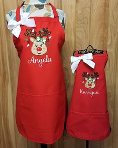 two red aprons with reindeer heads on them, one is personalized and the other has an embroidered name