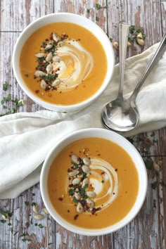 two bowls of carrot soup on a wooden table with silver spoons and napkin next to them